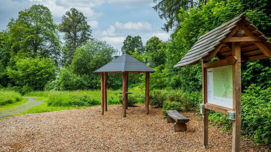 Eingang eines FriedWalds mit Infotafel, Sitzbank und überdachtem Holztisch – umgeben von grüner Natur und Waldweg.