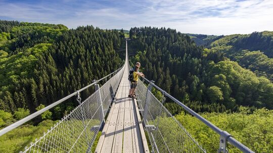Wanderer steht auf der Hängeseilbrücke Geierlay, die über ein bewaldetes Tal inmitten grüner Hügel führt.