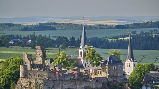 Blick auf die Kastellauner Burg mit Ruinen, Kirchen und Fachwerkhäusern inmitten einer weiten, grünen Hügellandschaft im Hunsrüc