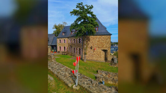 Historisches Burggemäuer mit Fachwerkfenstern und rot-weiß gestreiften Fensterläden, im Vordergrund ein Kunstobjekt mit roten Fl