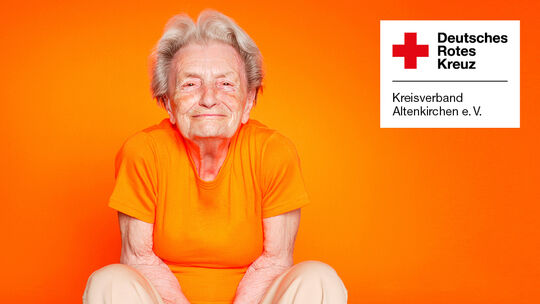 Ältere Frau in Orange vor DRK-Logo auf orangefarbenem Hintergrund