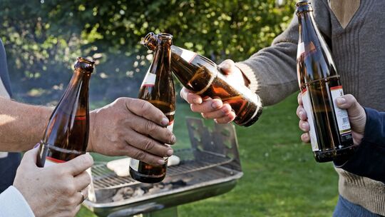 Vier Leute stoßen draußen vor einem Grill mit Bierflaschen an