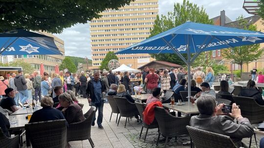 ahlreiche Menschen sitzen und stehen auf einem Platz mit Sonnenschirmen, entspannte Atmosphäre bei einem Fest.