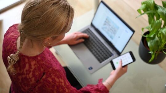 Frau sitzt vor einem Laptop und schaut auf ein Smartphone