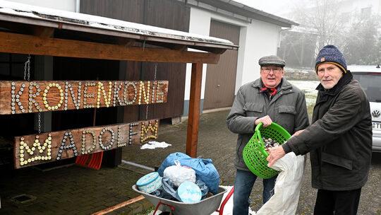 Adolf Schuch und Bernhard Helmich füllen vor dem auf Holzbohlen gefertigten Schild „Kronenkönig Adolf“ die gesammelten Kronkorke