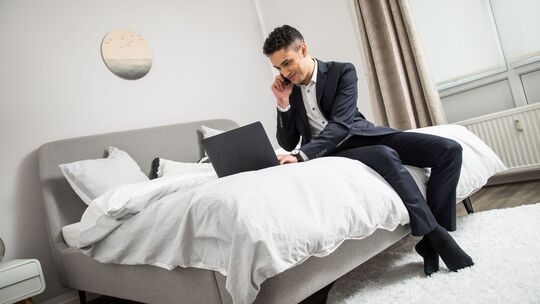 Ein Mann im Anzug sitzt auf einem Bett und arbeitet am Laptop Ein Mann im Anzug sitzt auf einem Bett und arbeitet am Laptop