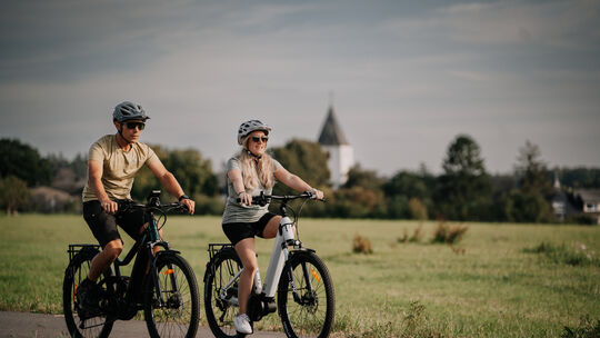 Zwei Personen fahren mit Fahrrädern über einen Radweg durch grüne Wiesen. Bildtitel: Wandern bei Kastellaun
