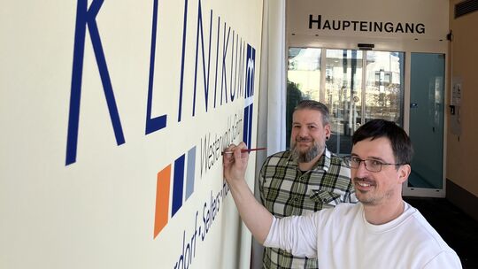 Matthias Dreser (rechts) hat den neuen Namen im Eingangsbereich des Selterser Krankenhauses an die Wand gemalt. Beim Anbringen d