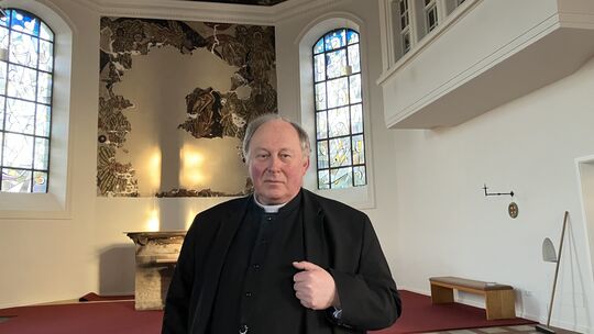 Dechant Martin Kürten steht dort, wo vor gut drei Jahren das Feuer brannte. Im Hintergrund das teilweise zerstörte Wandfresko En