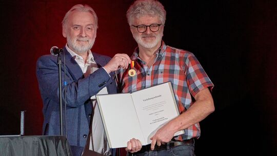 Kulturstaatssekretär Jürgen Hardeck überreicht Uli Schmidt die Landesverdienstmedaille für sein "jahrzehntelanges Engagement für