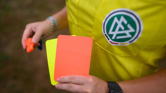 Eine Schiedsrichterin, steht mit roter und gelber Karte auf einem Rasen und hält in der anderen Hand eine Trillerpfeife. 961 Mal