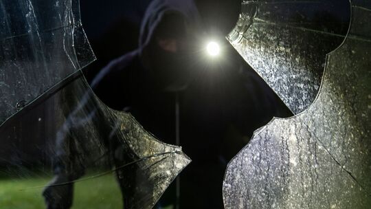 Durch eine eingeschlagene Scheibe blickt ein fiktiver Einbrecher