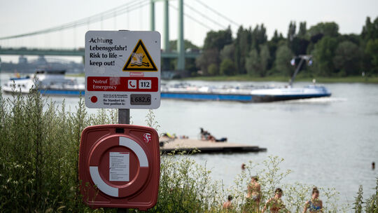Menschen sonnen sich am Rhein, während ein Frachtschiff vorbei fährt. Behörden und die Deutsche Lebens-Rettungs-Gesellschaft (DL