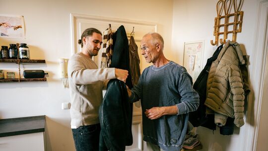 Ein junger Mann hilft einem Senior die Jacke anzuziehen Ein junger Mann hilft einem Senior die Jacke anzuziehen