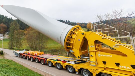 Ein Flügel eines Windrads ist auf einem sogenannten Selbstfahrer montiert und wird durch einen Ort transportiert.