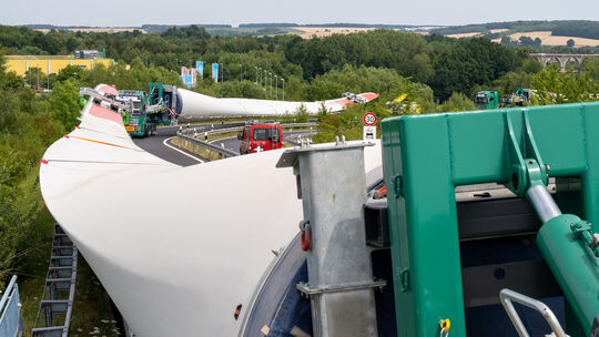 Ein Schwerlasttransport ist mit drei Lkws und aufgeladenen Flügeln für eine Windkraftanlage unterwegs.