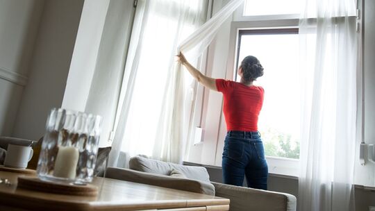 Eine Frau zieht eine Gardine vor einem gekippten Fenster auf