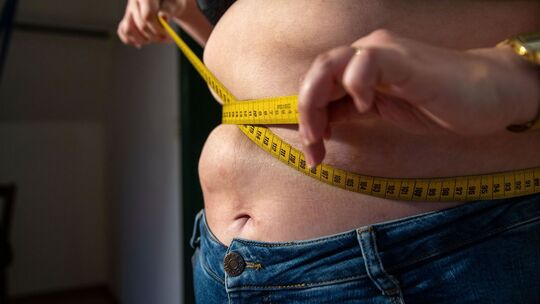 Eine Frau mit Übergewicht hält ein Maßband um ihren Bauch.
