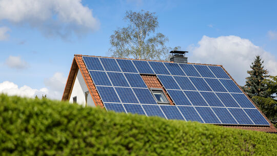 Ein Einfamilienhaus mit Solarpaneelen auf dem Hausdach steht hinter einer grünen Hecke. Immer mehr Unternehmen und Privathaushal