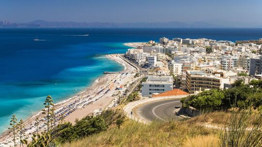 Blick auf die Küstenstadt Rhodos mit türkisblauem Meer, langem Sandstrand und Bergen im Hintergrund.